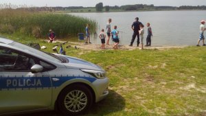 Patrol Policji nad brzegiem jeziora, z przodu stoi radiowóz, w oddali policjanci rozmawiają z dziećmi nad brzegiem jeziora.