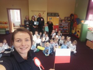 selfie zrobione przez st. sierż. Martę Szulc, w tle dzieci i wychowawcy, dzielnicowa trzyma chorągiewkę z barwami flagi polski