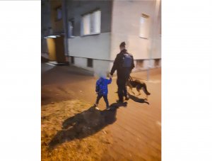 Policjant idzie chodnikiem obok bloku, za rękę trzyma chłopca.