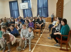 fotografia kilkudziesięciorga dzieci siedzących na sali ujęcie z boku sali, widać siedzących na bocznej ławce kilkoro nauczycieli