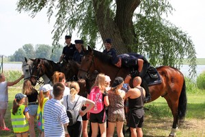 Policjanci na koniach stoją pod wierzbami, wraz z nimi są dzieci.