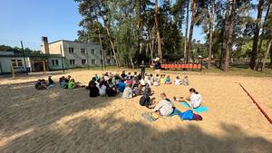 grupa dzieci siedząca na piasku na boisku słuchają wykładu prowadzonego przez profilaktyka społecznego