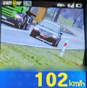 Widok monitora urządzenia pomiarowego, na którym widnieje pojazd z poruszający się z prędkością 102 km/h, dodatkowo pojazd  wyprzedza samochód ciężarowy i przekracza linię podwójną ciągłą
