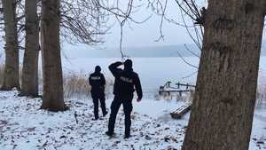policjanci podczas kontroli zbiorników wodnych w oddali widać dwóch policjantów wpatrujących sie w tafle zamarzniętego jeziora