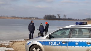 Na zdjęciu widać radiowóz, dwójkę dzieci rozmawiającą z policjantem, w tle jest zamarznięte jezioro.