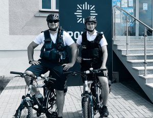 Policjanci siedzą na rowerach, w tle znak z logotypem POLICJA