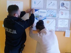 policjant zawiesza wraz z nauczycielem plakat dotyczący usług wielkopolskiej Policji