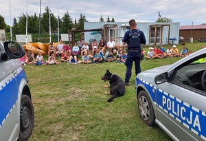 Policjant przewodnik psa służbowego stoi przed dziećmi , które go słuchają siedząc na trawniku