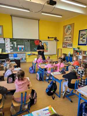 Policjantka prowadzi zajęcia dla dzieci. Stoi w klasie, dzieci siedzą w ławkach.