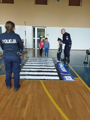 Policjanci uczą dzieci prawidłowego przejścia przez przejście dla pieszych. Ćwiczenia na sali przy rozkładanej dydaktycznej zebrze