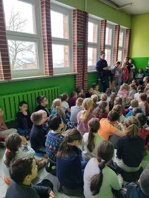Dzieci zgromadzone na dużym korytarzu słuchają prelekcji policjantki dzielnicowej