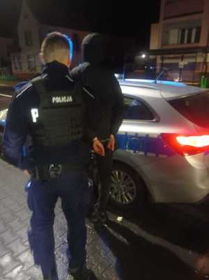 jest noc, policjant stoi z zatrzymanym mężczyzną, który ma zakute ręce, stoją tyłem przy radiowozie