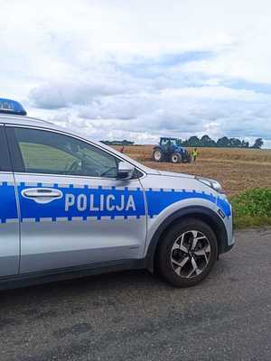 fotografia przedstawia radiowóz i w tle na polu ciągnik rolniczy