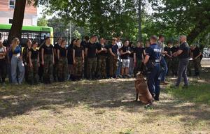 Policjant z psem stoi przed grupą uczniów na trawniku i prowadzi zajęcia