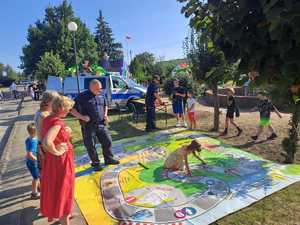 policjant prowadzi grę edukacyjną dla dzieci. Plansza po której uczestnicy mogą chodzić, jest rozłożona na trawniku