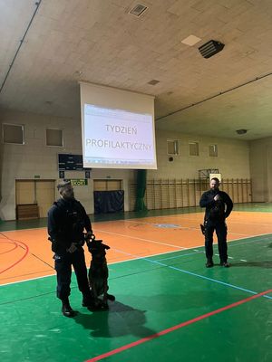 policjant  z psem stoi, pies obok niego siedzi na parkiecie hali