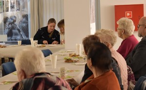 policjantka siedząc rozmawia z kobieta obok, przy stołach siedzą seniorzy.