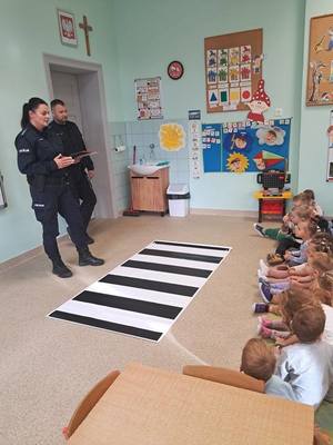 policjanci stoją ze znakami przed symulatorem przejścia dla pieszych, w klasie siedzą dzieci