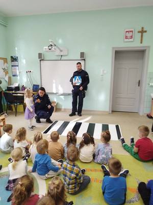 policjanci stoją ze znakami przed symulatorem przejścia dla pieszych, w klasie siedzą dzieci, dziecko pokazuje jak przejść