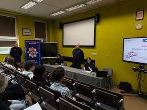 zdjęcie przedstawia policjantów prowadzących spotkanie, w tle tablica z monitorem i prezentacją, w ławkach siedzą słuchacze, prelegenci prowadzą zajęcia