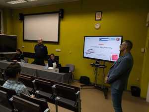 zdjęcie przedstawia policjantów prowadzących spotkanie, w tle tablica z monitorem i prezentacją, w ławkach siedzą słuchacze, prelegenci prowadzą zajęcia