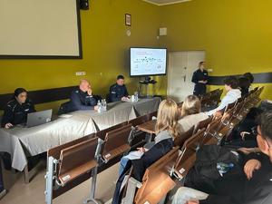 zdjęcie przedstawia policjantów prowadzących spotkanie, w tle tablica z monitorem i prezentacją, w ławkach siedzą słuchacze, prelegenci prowadzą zajęcia