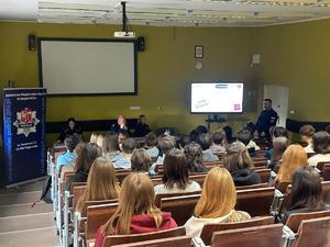 zdjęcie przedstawia policjantów prowadzących spotkanie, w tle tablica z monitorem i prezentacją, w ławkach siedzą słuchacze, prelegenci prowadzą zajęcia