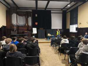 policjant podczas prowadzenia zajęć w auli szkoły, nosi kamizelkę z napisem technik kryminalistyki, na zdjęciu również widoczna walizka na zdarzenia, do tego przybory wyłożone na stole.